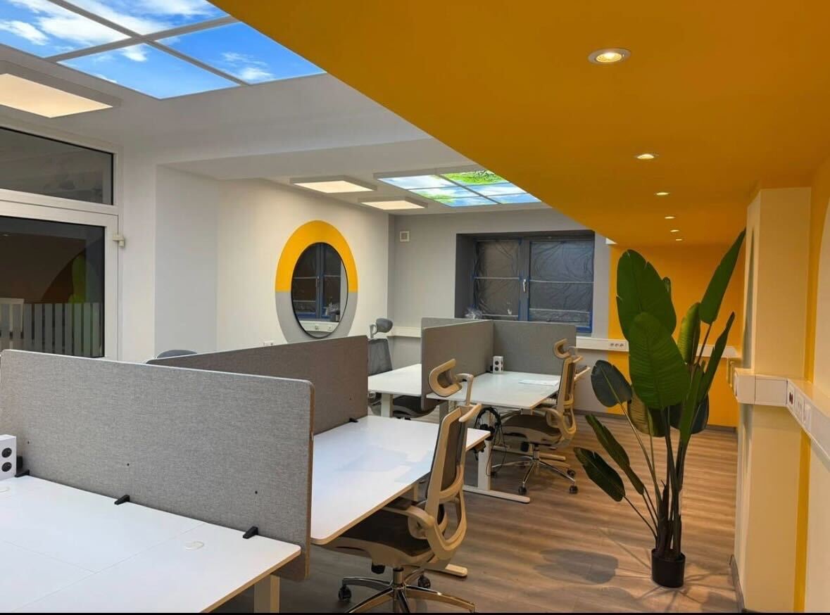 Coworking Space CoWork Station in Bakede – Arbeitsplätze im Open Space Coworking Space CoWork Station in Bakede bei Bad Münder: helle Arbeitsplätze mit weißen Tischen, grauen Sichtschutz-Trennwänden, ergonomischen Stühlen, gelber Akzentwand und Himmeldecken-Panel mit Wolkenmotiv